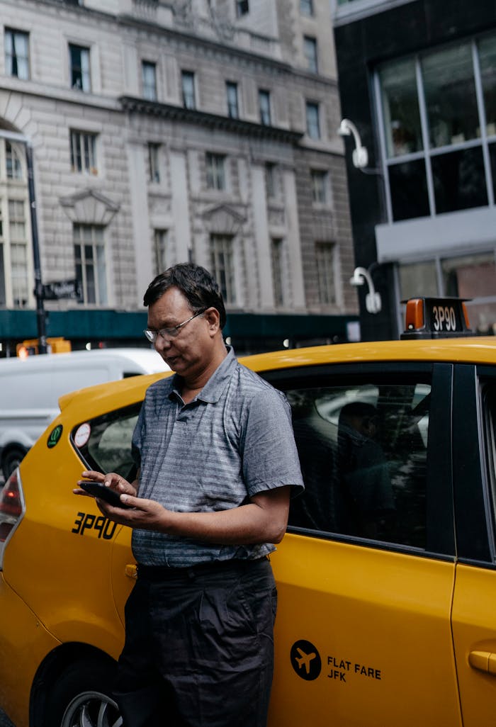 A taxi driver standing outside a yellow cab, using a smartphone on a busy city street.